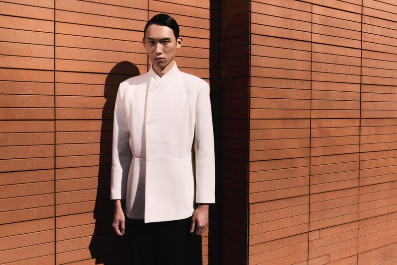 East Asian male model posing in an off-white structured high-neck jacket and black trousers against a terracotta tile wall.