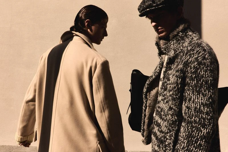 Two men in fashionable winter coats stand against a light wall, one in a beige coat, the other in a black-and-white knit coat.