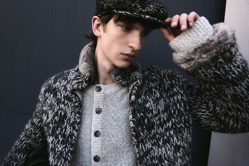 Portrait of a young adult male wearing a newsboy cap and wool coat, posing in a studio with a dark background.