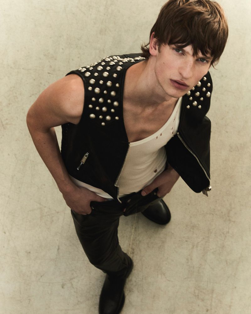 Man in studded vest poses with hands in his pockets, looking at the camera from a high angle in a fashion-editorial style shot.