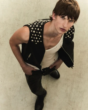 Man in studded vest poses with hands in his pockets, looking at the camera from a high angle in a fashion-editorial style shot.