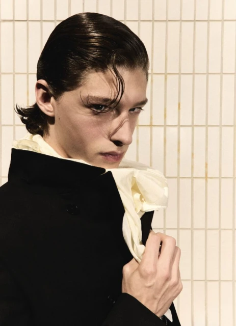Close-up portrait of a young adult male model with slicked-back hair wearing a black coat with a white ruffled collar, against a neutral background.