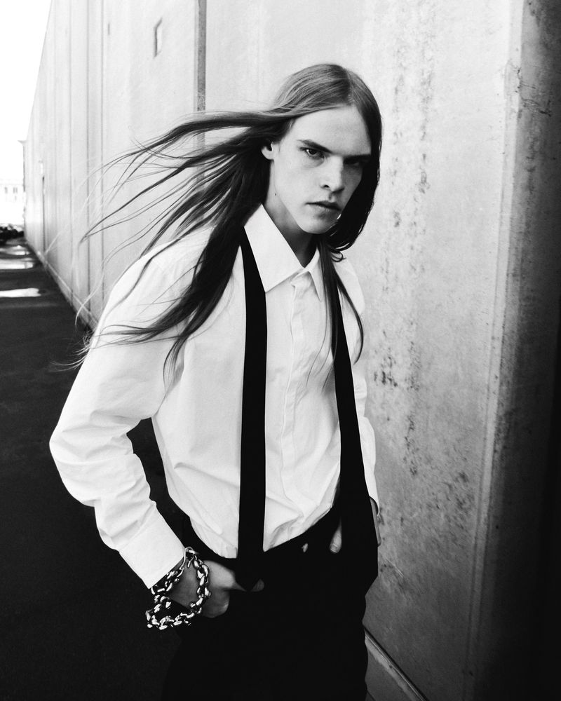 Black and white portrait of a young adult male with long hair, wearing a white shirt and suspenders, posing outdoors in an urban setting.
