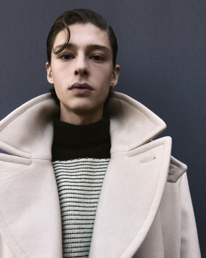 Headshot of a young adult male with slicked-back dark hair, wearing a cream-colored coat over a striped sweater, looking directly at the camera.
