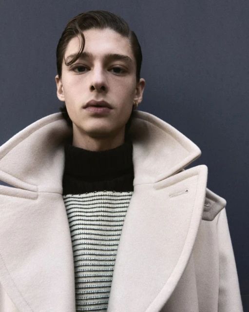 Headshot of a young adult male with slicked-back dark hair, wearing a cream-colored coat over a striped sweater, looking directly at the camera.