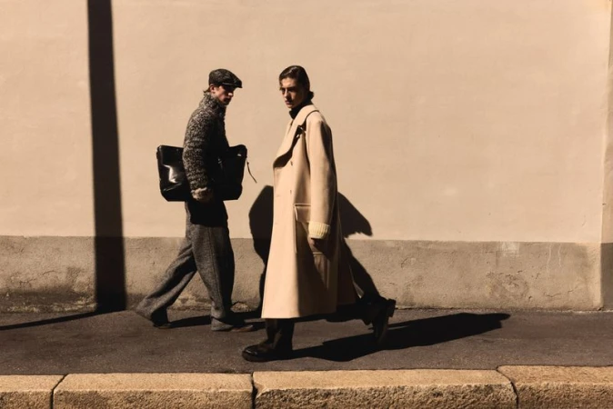 Two men walking past a textured wall, one wearing a long beige coat and the other a tweed cap and patterned sweater holding a black bag.