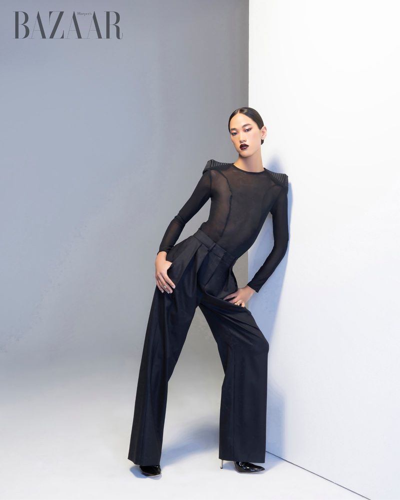 A model poses in a dark bodysuit and high-waisted, wide-legged trousers leaning against a white wall in a fashion editorial studio shot.
