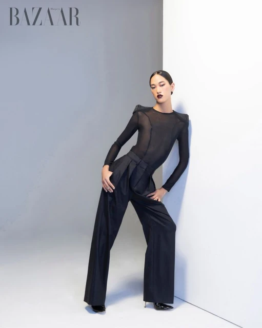 A model poses in a dark bodysuit and high-waisted, wide-legged trousers leaning against a white wall in a fashion editorial studio shot.