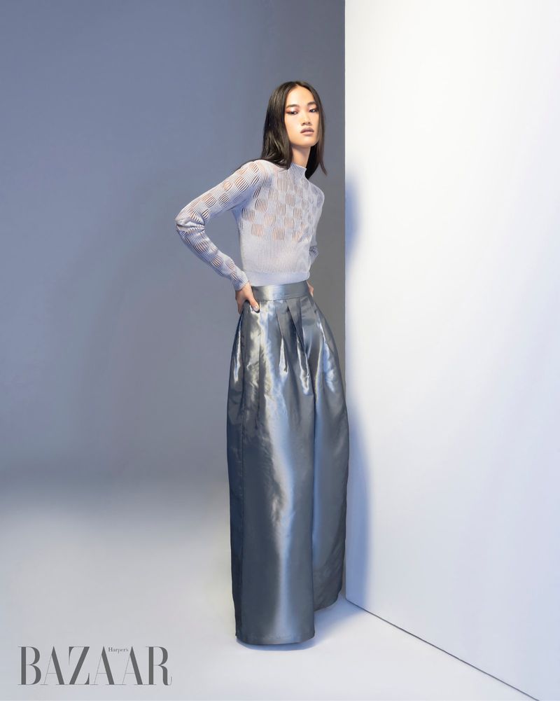 Model Parker Foubert poses in a sheer patterned knit top and metallic silver wide-leg trousers against a minimal studio wall.