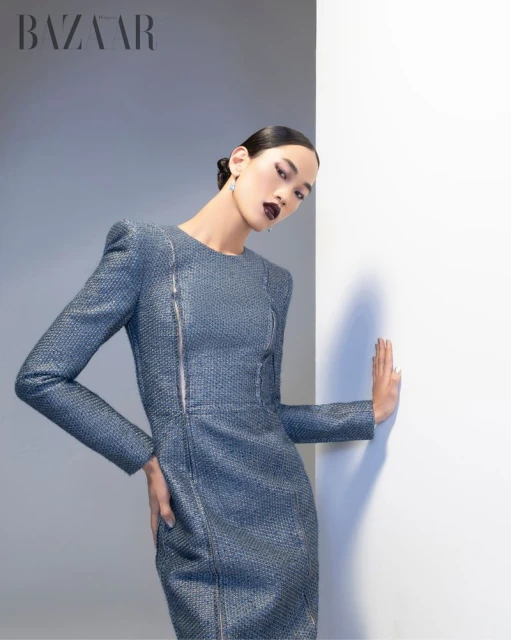 An Asian woman poses in a textured grey dress with a zipper for a fashion editorial photo, hands resting on a white wall in studio lighting.