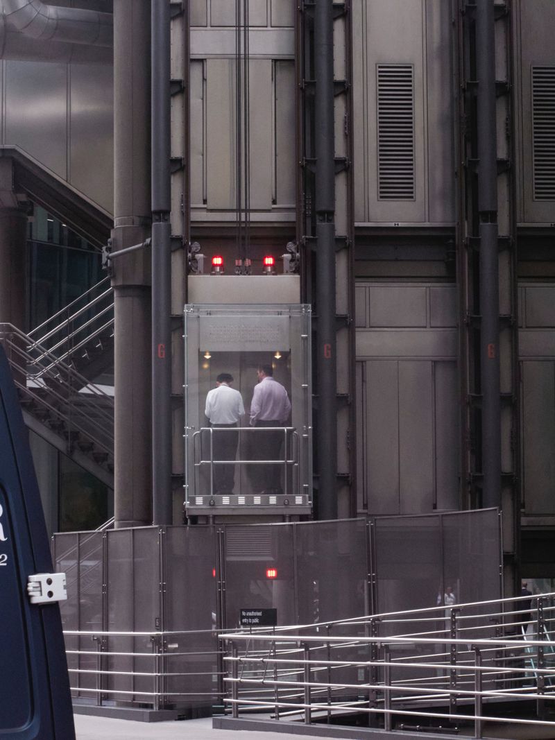 Two men inside a glass elevator in a building with exposed pipes and structural elements, ascending within an urban environment.