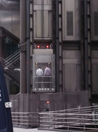 Two men inside a glass elevator in a building with exposed pipes and structural elements, ascending within an urban environment.