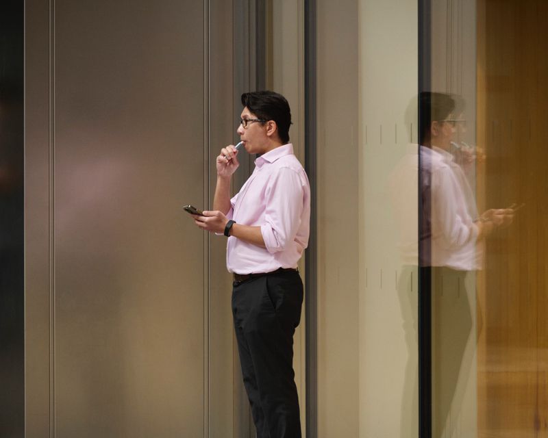 Man in a light pink button-down shirt and black trousers holding a smartphone in front of a modern glass office facade.