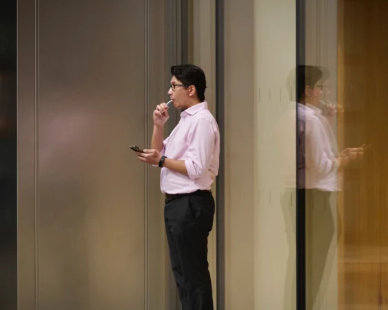Man in a light pink button-down shirt and black trousers holding a smartphone in front of a modern glass office facade.