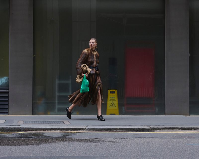 A woman in a fur coat walks on a city street carrying a bag, with a yellow caution sign visible in the background on a cloudy day.