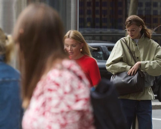 A woman with glasses and a bag walks down the street, accompanied by other pedestrians and city buildings in the background.