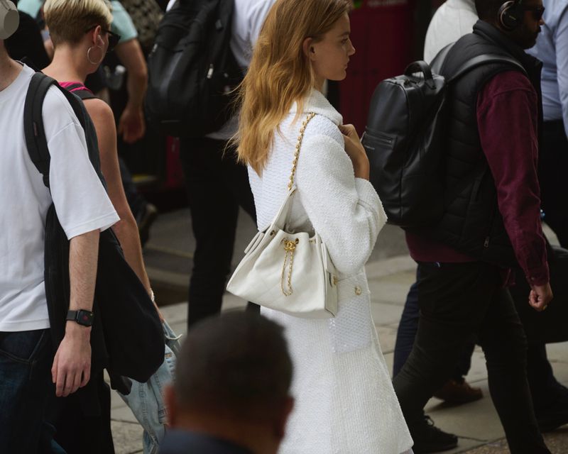 A woman walks in a busy urban environment carrying a Chanel handbag, wearing a white coat, with other people around her.
