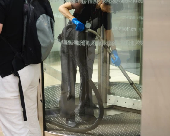 Adult female in black uniform and blue gloves is vacuuming the floor by a revolving glass door. A man in white pants stands to her side.