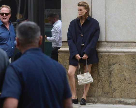 Model standing against a stone pillar wearing an oversized dark coat and holding a white handbag.
