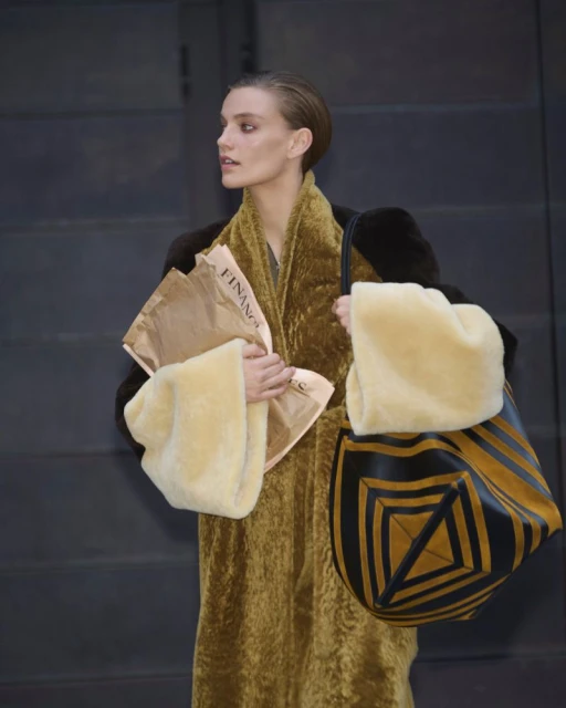A stylish woman in a coat carries a patterned bag and newspaper against a plain wall, showcasing a blend of fashion and texture.