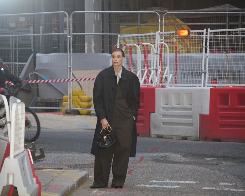 Woman in oversized black coat and olive suit stands on urban street with construction barriers in the background, holding black purse.