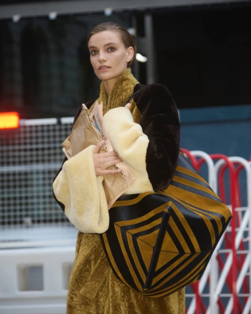 Fashionable woman in a textured coat carries newspapers and a large geometric tote bag in an outdoor, urban environment.