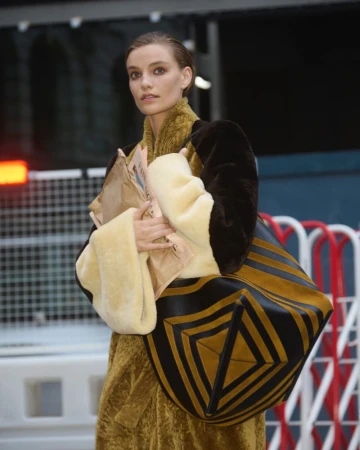 Fashionable woman in a textured coat carries newspapers and a large geometric tote bag in an outdoor, urban environment.