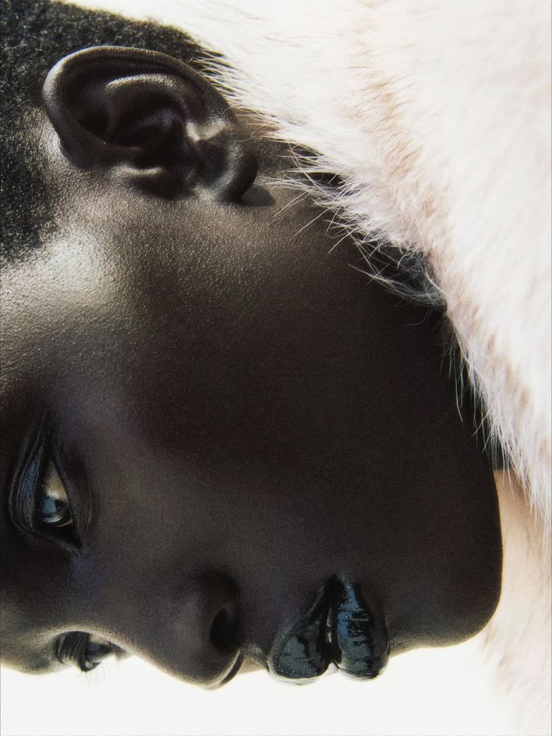 Close-up portrait of an African American woman with dark skin and glossy lips, adorned with a white furry collar against a light backdrop.