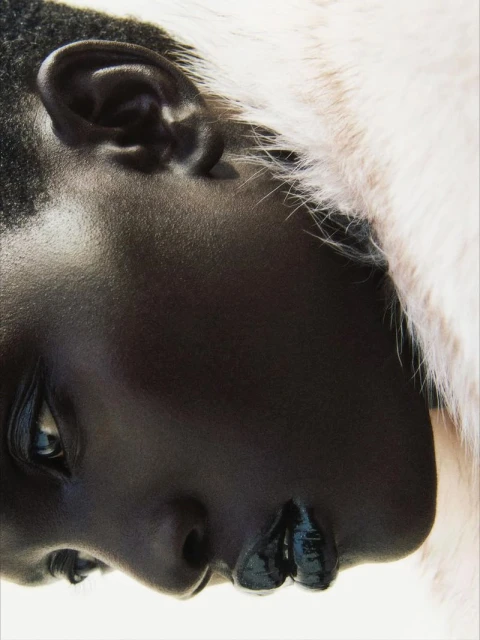 Close-up portrait of an African American woman with dark skin and glossy lips, adorned with a white furry collar against a light backdrop.