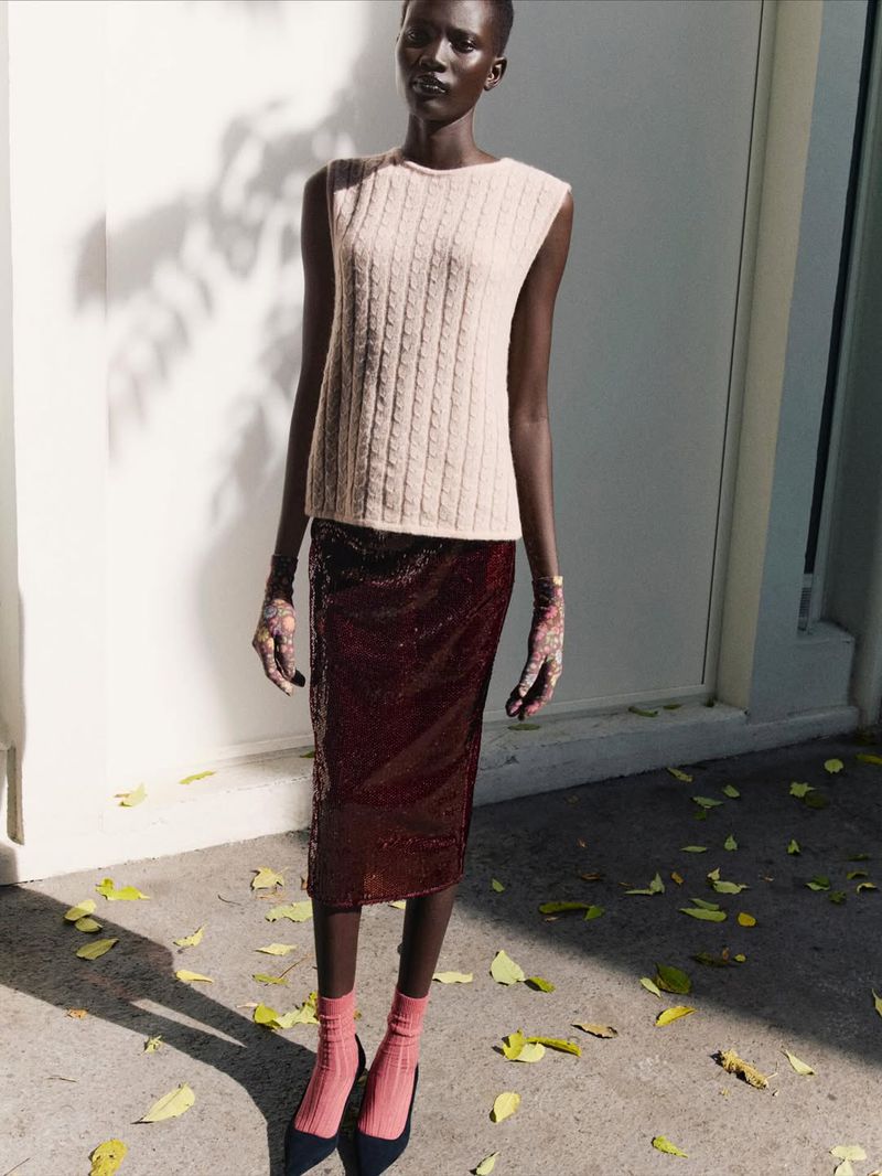 Fashion editorial shot of a Black female model wearing a cable-knit sweater, burgundy skirt, gloves, socks, and heels standing outdoors.