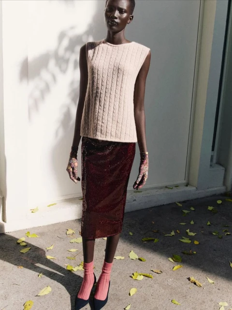 Fashion editorial shot of a Black female model wearing a cable-knit sweater, burgundy skirt, gloves, socks, and heels standing outdoors.