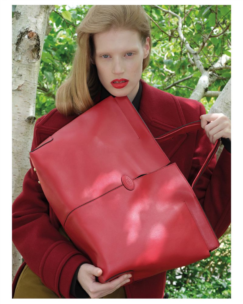 A fair-skinned adult woman with red lipstick is holding a large red leather bag outside, wearing a red wool coat against a blurred, leafy background.