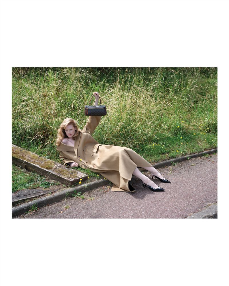 A woman in a long tan trench coat lies on a path holding a handbag in an outdoor fashion editorial, with tall green grass behind her.