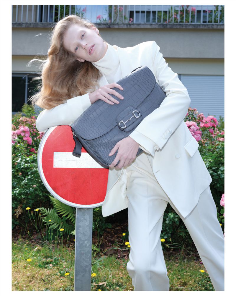 Fashion shot of a long-haired woman in an off-white suit holding a gray reptile-texture bag near a red and white no-entry sign.