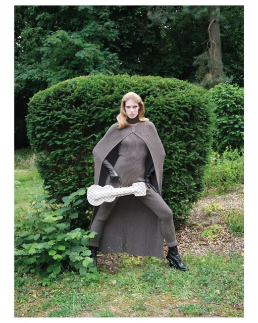 A long-haired female model poses in the park wearing a poncho and matching knitted suit, holding a guitar-shaped handbag.
