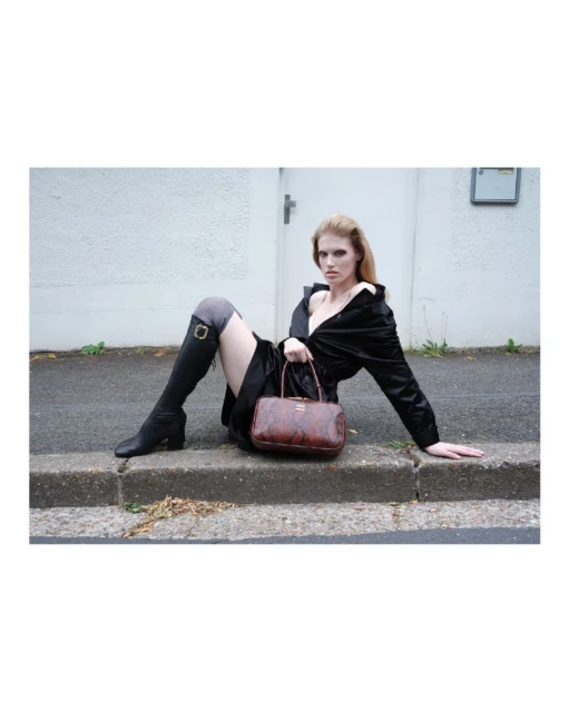 Fashion shot of a woman in black jacket and boots sitting against a white wall, holding a brown leather handbag with a snake pattern.