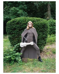 Model Jacqui Hooper poses in a taupe knit cape and jumpsuit holding a checkered guitar bag in front of a lush green garden hedge.