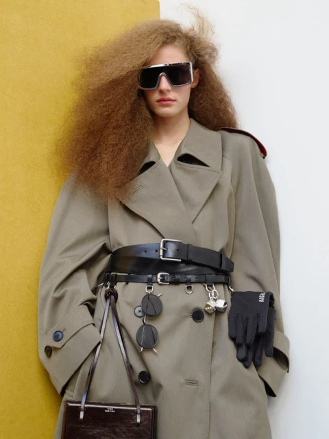 Fashionable woman with curly hair wears oversized sunglasses, tan trench coat, wide belt, and gloves against a mustard and white background.
