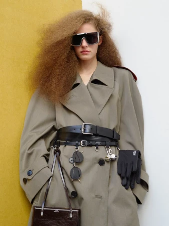 Fashionable woman with curly hair wears oversized sunglasses, tan trench coat, wide belt, and gloves against a mustard and white background.