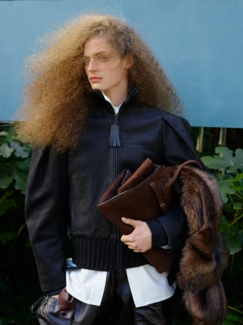 A woman with voluminous curly hair poses in a black leather jacket, holding a brown leather tote bag in an outdoor setting.