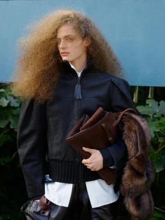A woman with voluminous curly hair poses in a black leather jacket, holding a brown leather tote bag in an outdoor setting.