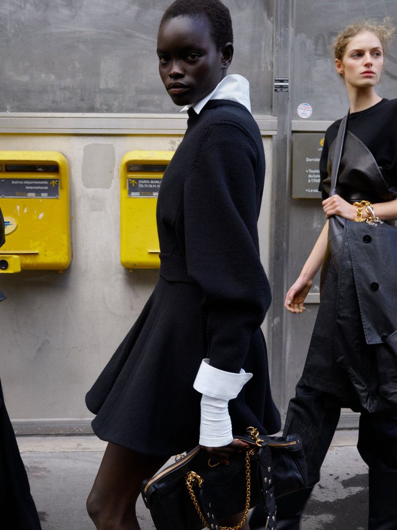 Fashion editorial shot of two women in stylish black dresses walking in an urban setting with yellow mailboxes in the background.