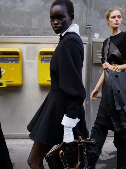Fashion editorial shot of two women in stylish black dresses walking in an urban setting with yellow mailboxes in the background.