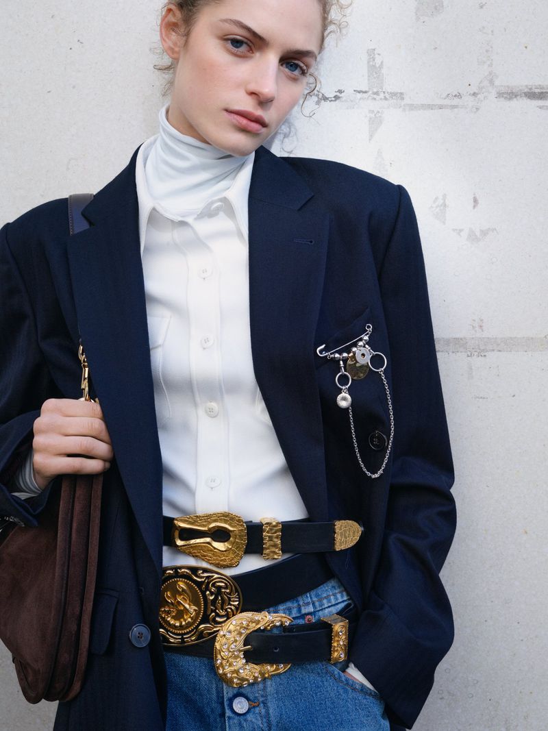 Fashionable woman in blazer and jeans accessorized with unique belts and a handbag, posing against a textured wall in soft, natural light.
