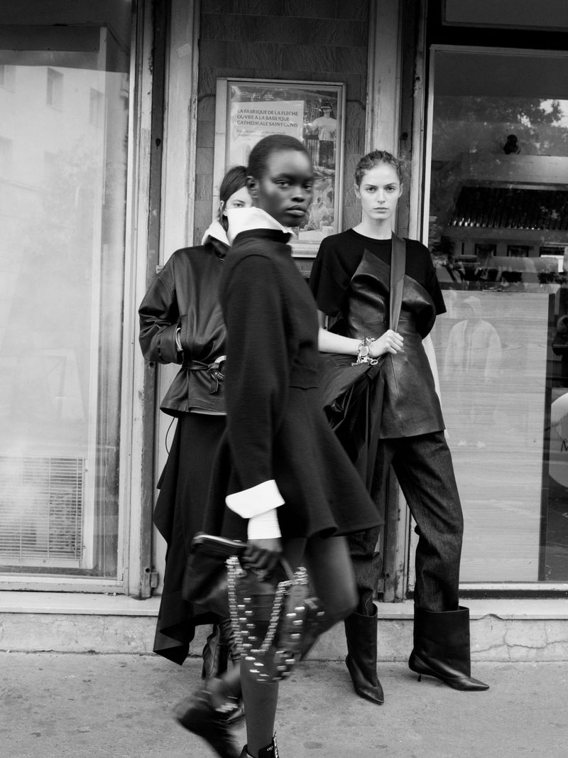 Three fashion models stand outside a store, wearing black outfits and holding a studded purse, captured in a monochromatic, medium shot.