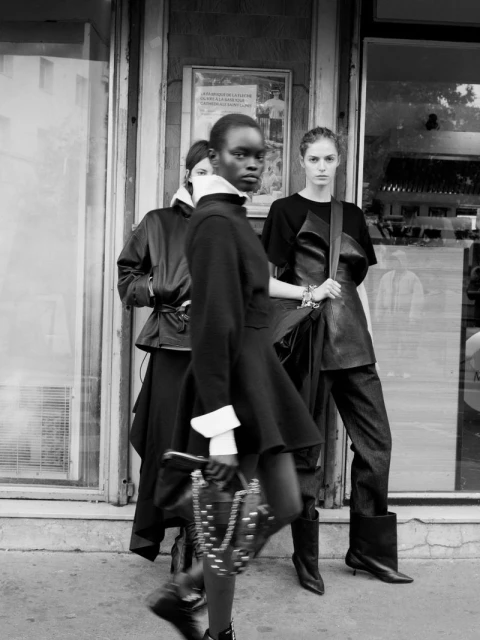 Three fashion models stand outside a store, wearing black outfits and holding a studded purse, captured in a monochromatic, medium shot.