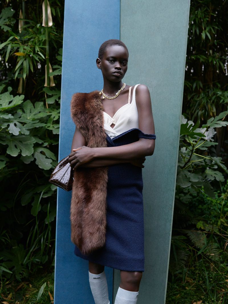 An elegant African woman poses in a garden wearing a navy dress, fur stole, jewelry, and knee socks in a fashion editorial shot.
