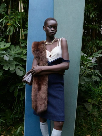 An elegant African woman poses in a garden wearing a navy dress, fur stole, jewelry, and knee socks in a fashion editorial shot.
