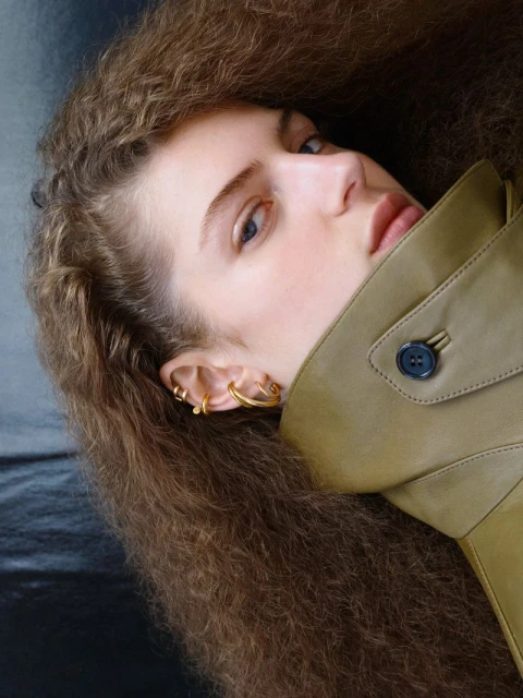 A young woman with curly brown hair and gold earrings, wearing a tan trench coat, poses in a close-up studio portrait with a black backdrop.