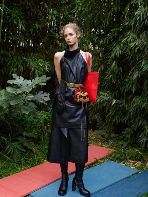 Fashionable adult female posing in black dress, boots, and yellow belt with red bag in natural outdoor setting.